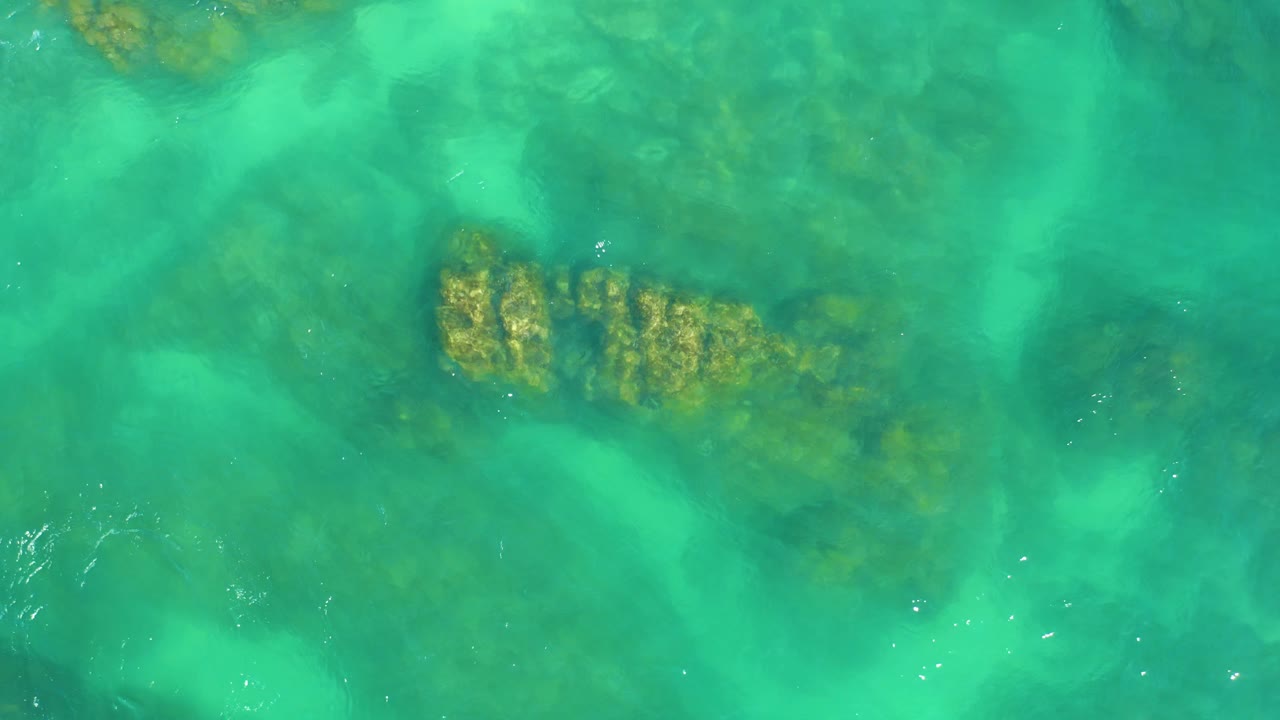 Shallow turquoise ocean water covers a darker seabed and a yellow-brown coral and rock cluster at Dreams Los Cabos, with no visible people.