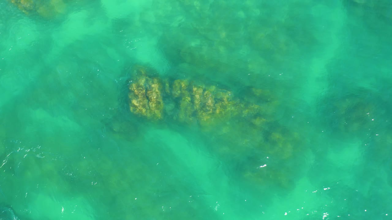Green-blue water runs over visible seabed rocks and coral near the lower left of the frame at Dreams Los Cabos, with no visible people.