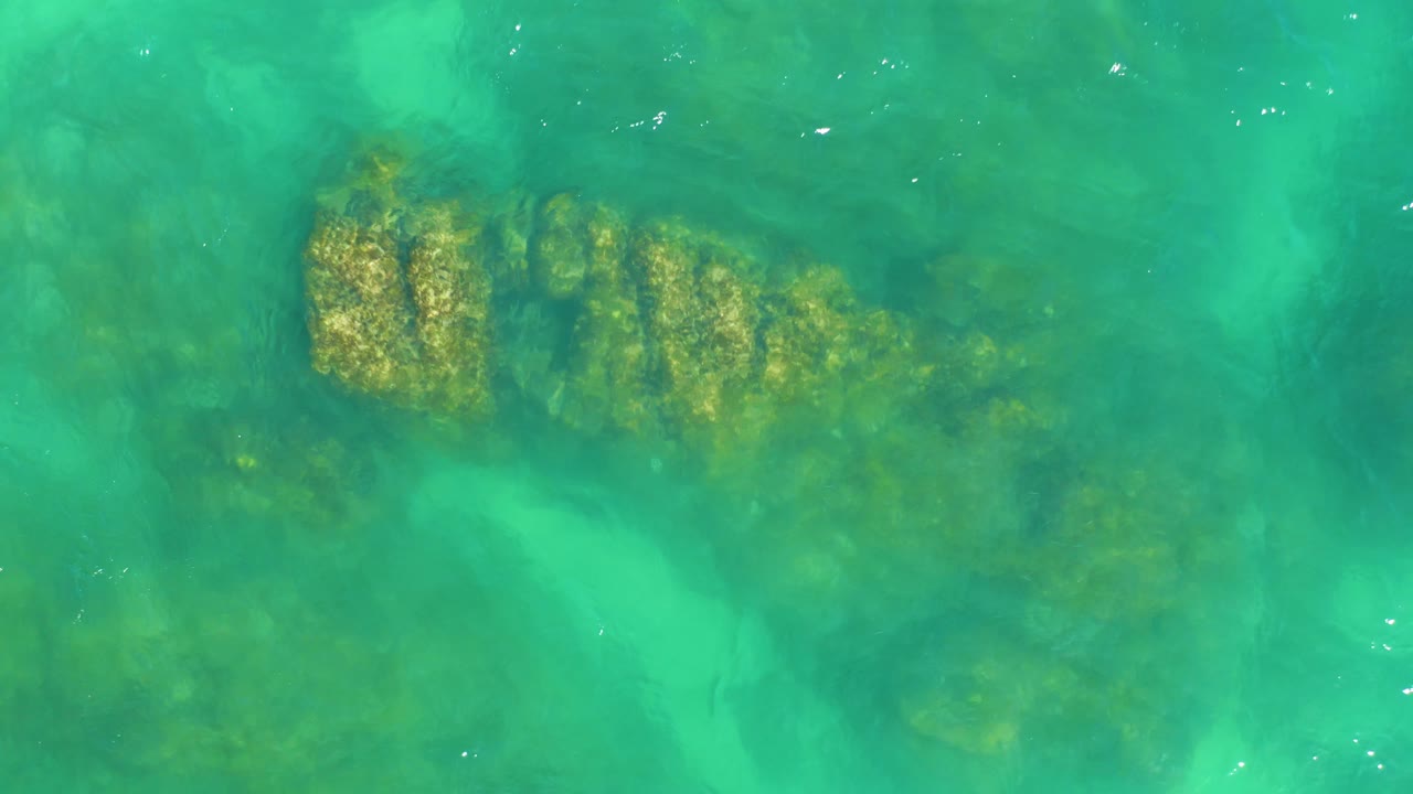 At Dreams Los Cabos, a rocky coral patch sits under turquoise water with brighter surface sparkle near the lower center, and no visible people.