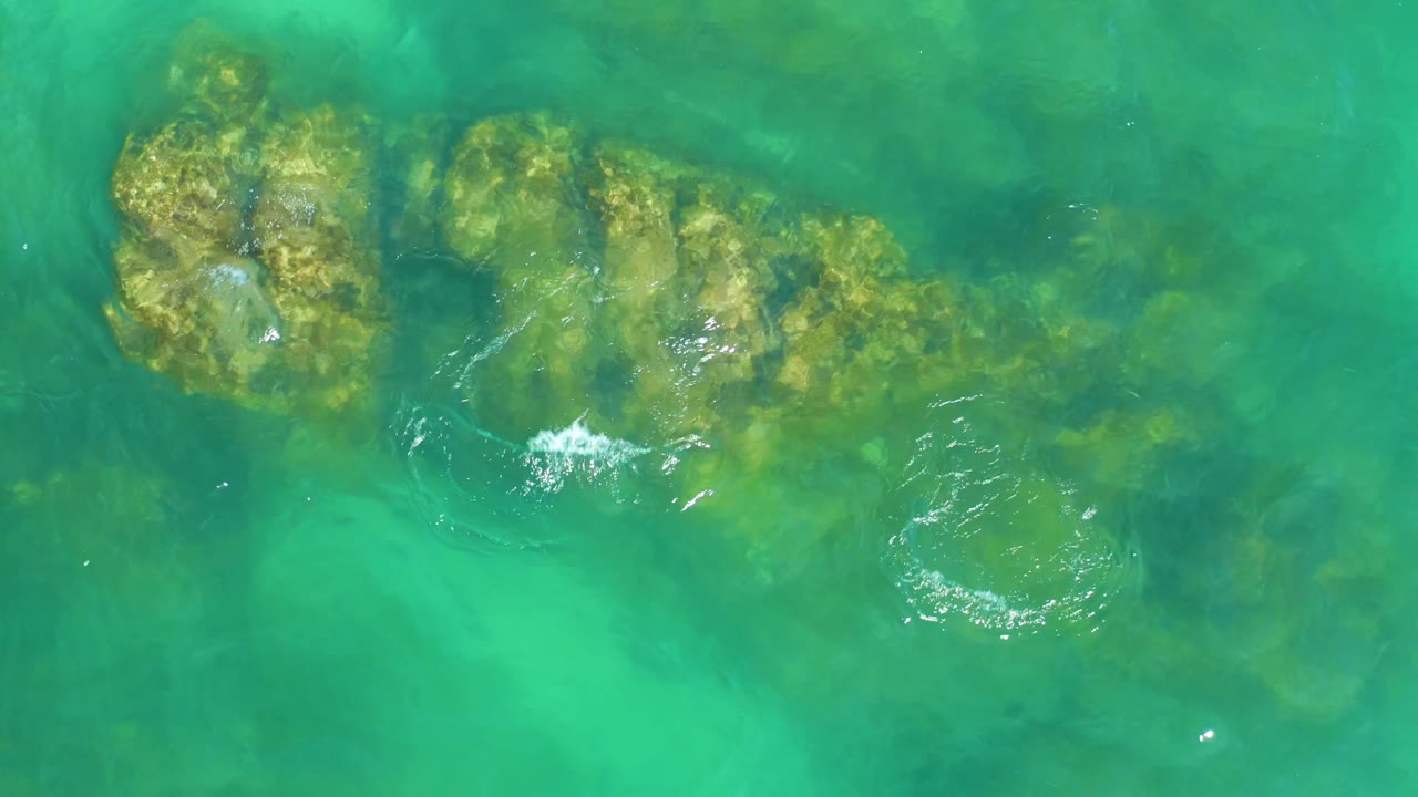 Aerial view at Dreams Los Cabos with the main submerged rock cluster centered under clear turquoise water, white ripple highlights, and no visible people.