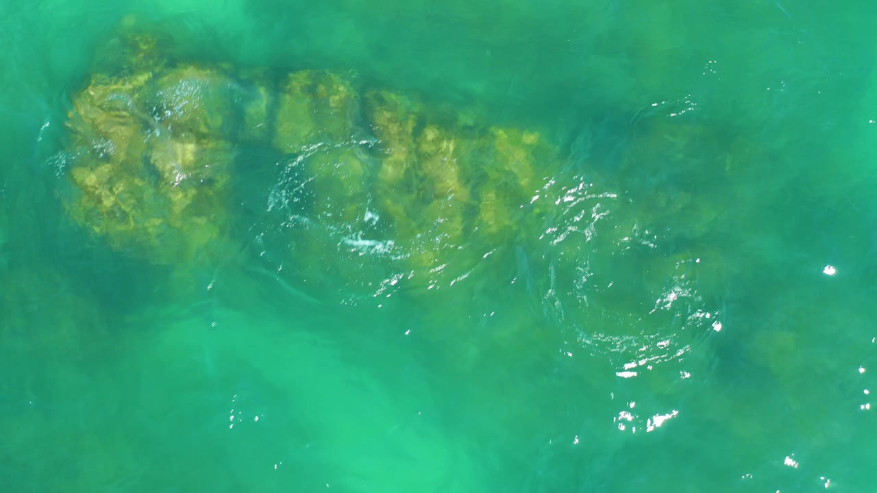 Overhead Dreams Los Cabos water detail with a long algae-tinted rock ledge under green-blue water and brighter reflections along the right side, with no visible people.