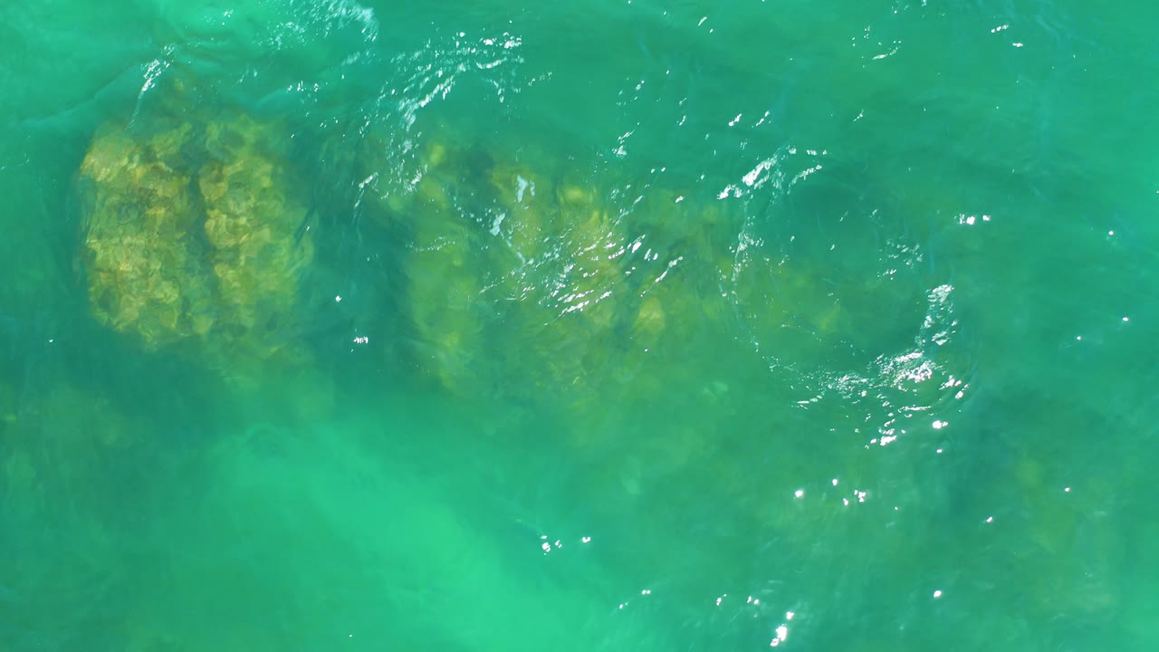Aerial frame from Dreams Los Cabos showing the left edge of a submerged rocky outcrop under transparent turquoise water, with ripples crossing open water and no visible people.