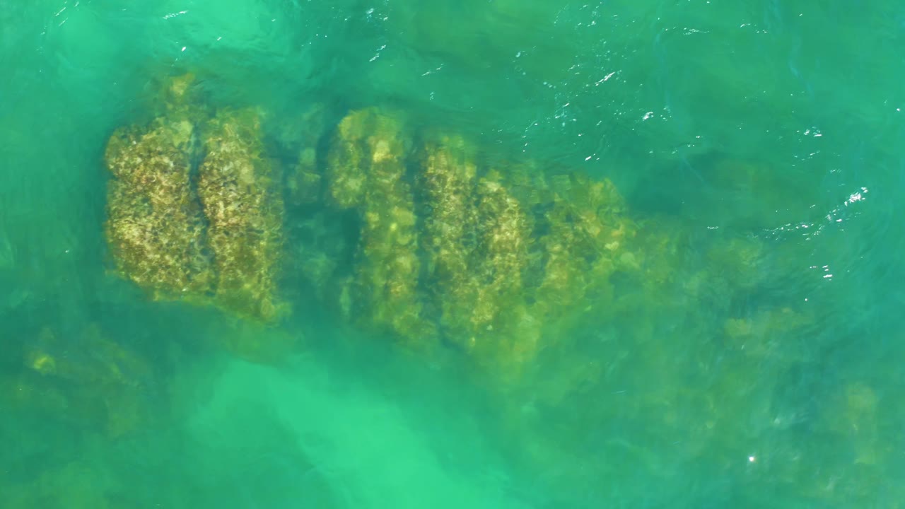 Overhead frame of clear green-blue water at Dreams Los Cabos with the submerged rock band centered left and no visible people.