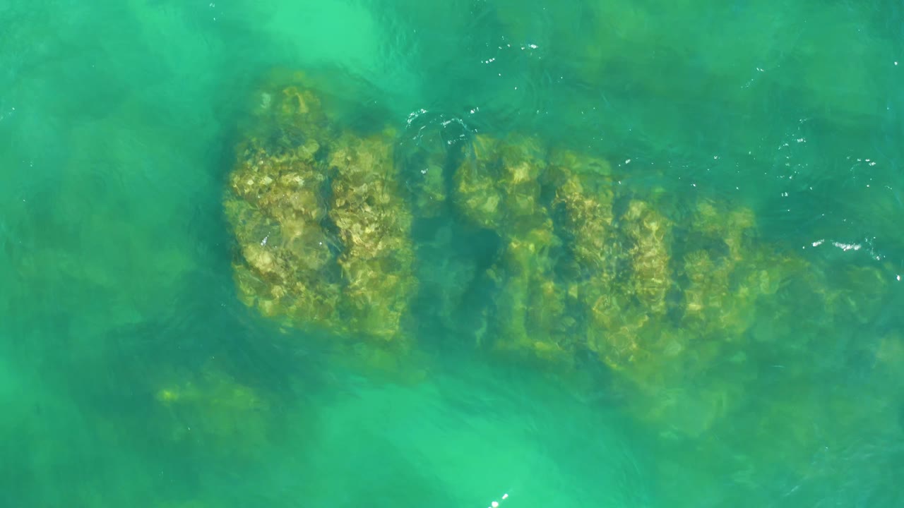 Clear green-blue water ripples over a long brown underwater rock formation at Dreams Los Cabos, with bright surface highlights and no visible people.