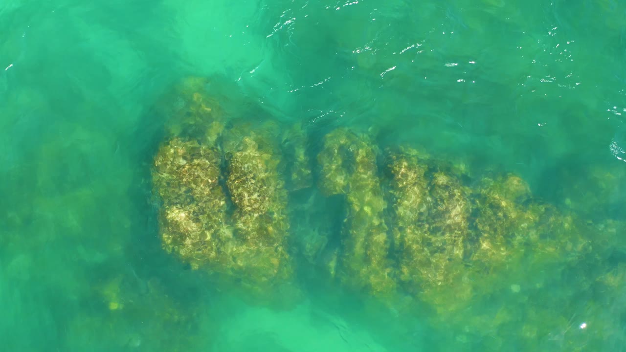 Clear water at Dreams Los Cabos reveals a central cluster of submerged rocks as small ripples soften the edges, with no visible people.