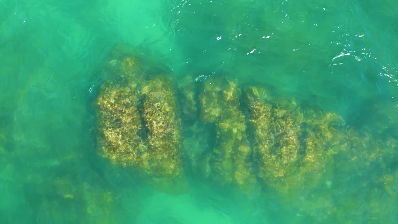 Turquoise ocean water at Dreams Los Cabos covers a long underwater rock formation, with bright flecks of surface reflection and no visible people.