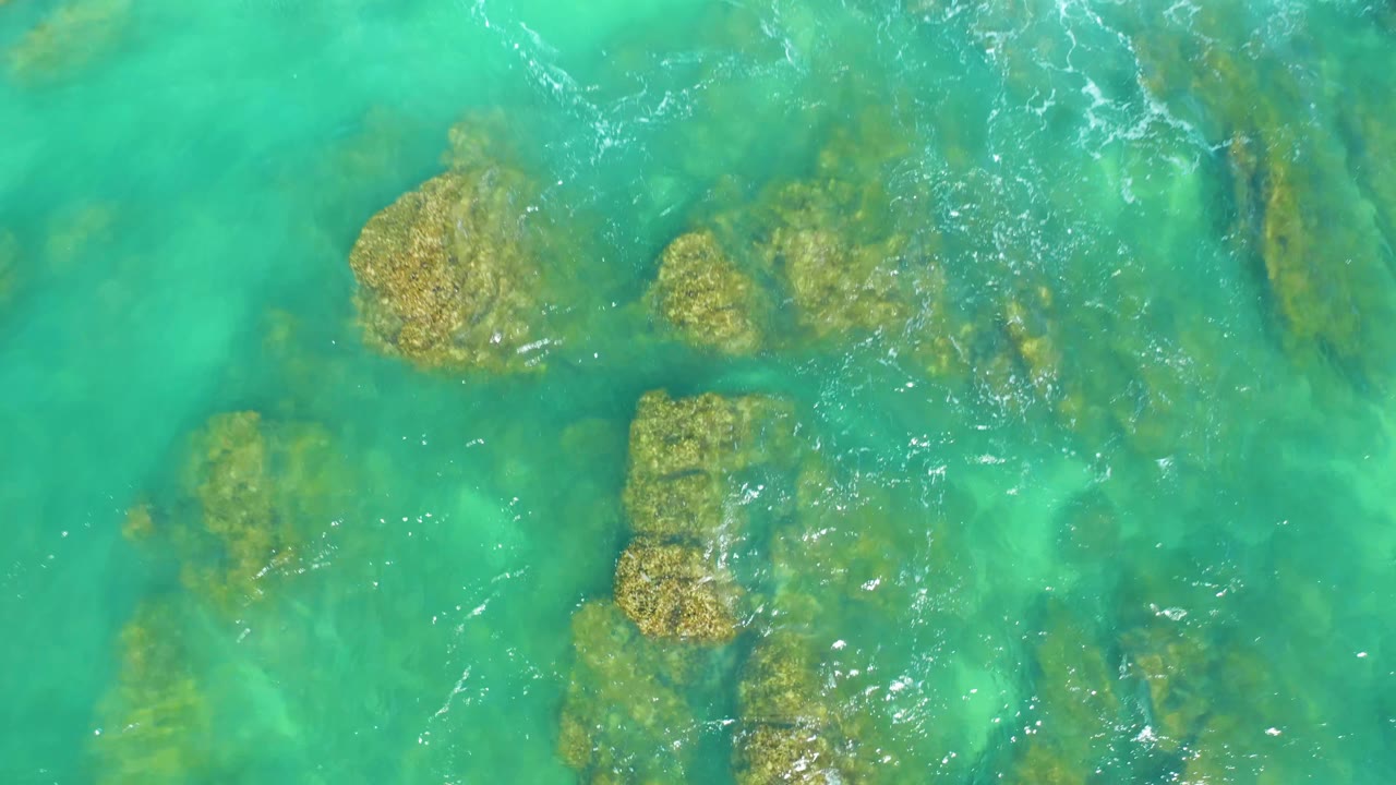 Clear shallow ocean water reveals clustered rocks beneath the surface at Dreams Los Cabos, with small ripples crossing the upper right and no visible people.