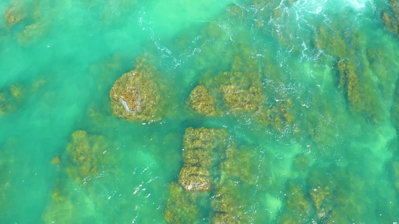 A foamy wave edge enters the upper right of the turquoise water above submerged rocks at Dreams Los Cabos, with no visible people.