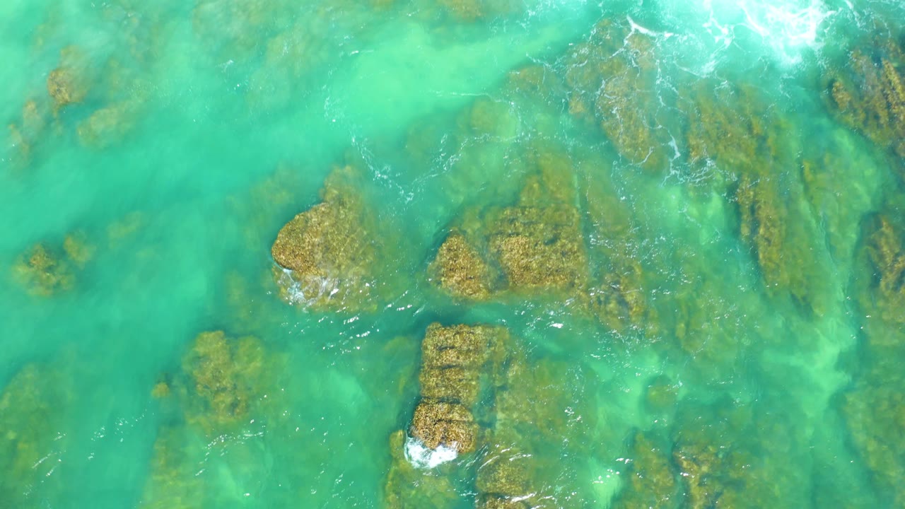 Green-blue shallow water covers a rocky seabed at Dreams Los Cabos, with several larger rock shapes near the lower half and no visible people.