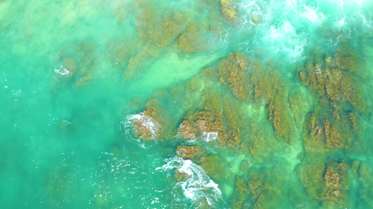 At 0:01, the Dreams Los Cabos overhead view shows green-blue water, clustered submerged rocks, and a brighter foam trail forming near the lower left with no visible people.