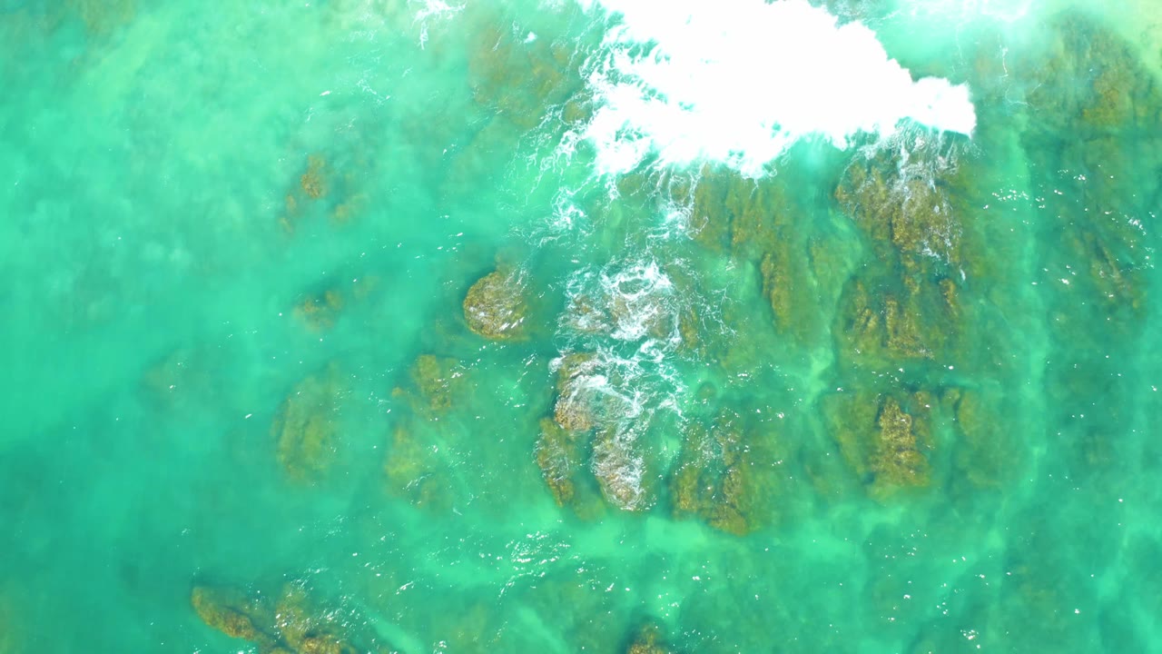 Clear blue-green water at Dreams Los Cabos reveals scattered underwater rocks while a white foam band sits near the upper frame, with no visible people.