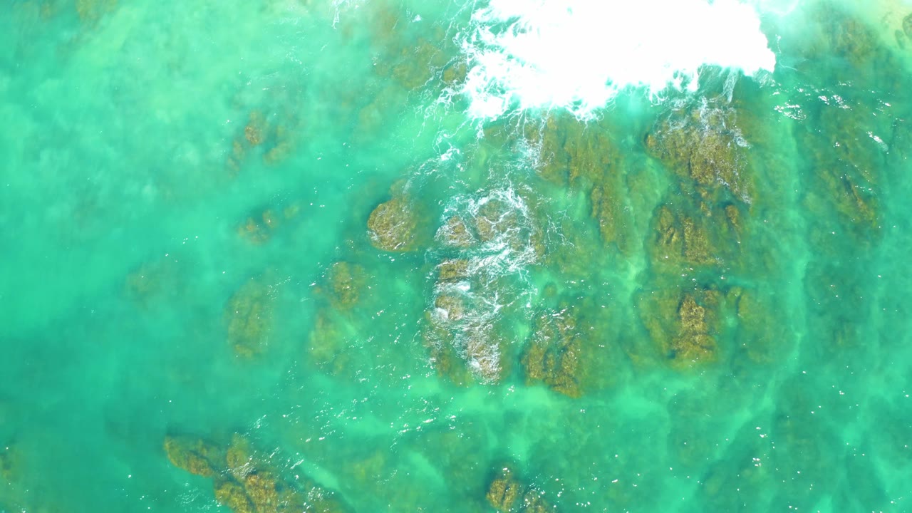 Top-down view at Dreams Los Cabos showing turquoise shallows, brown submerged rocks, a bright foam patch along the top, and no visible people.
