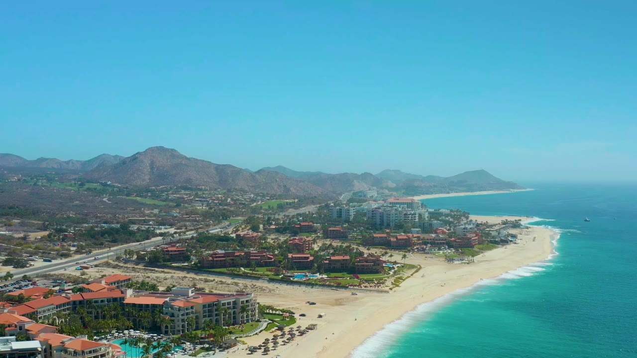 At 0:00, an aerial view over Dreams Los Cabos shows red-roofed resort buildings, a wide sandy beach, turquoise water, and mountains under a clear sky.