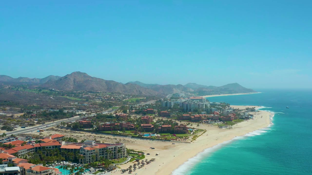 Wide aerial coastline with resort roofs, beach umbrellas, surf, and mountains.