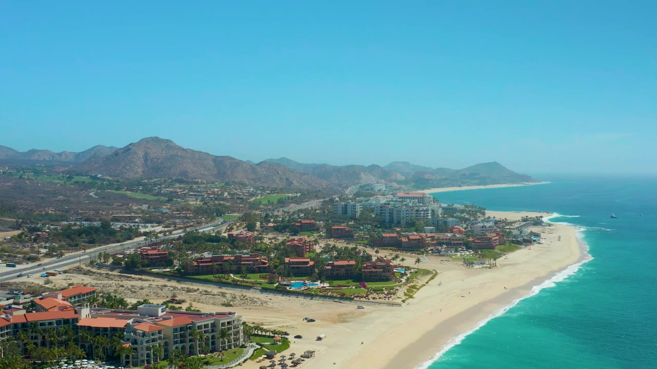 Aerial view at 0:01 over Dreams Los Cabos, showing the highway and small vehicles to the left, beachfront resort buildings in the center, pale sand and blue-green water to the right, and no visible people.