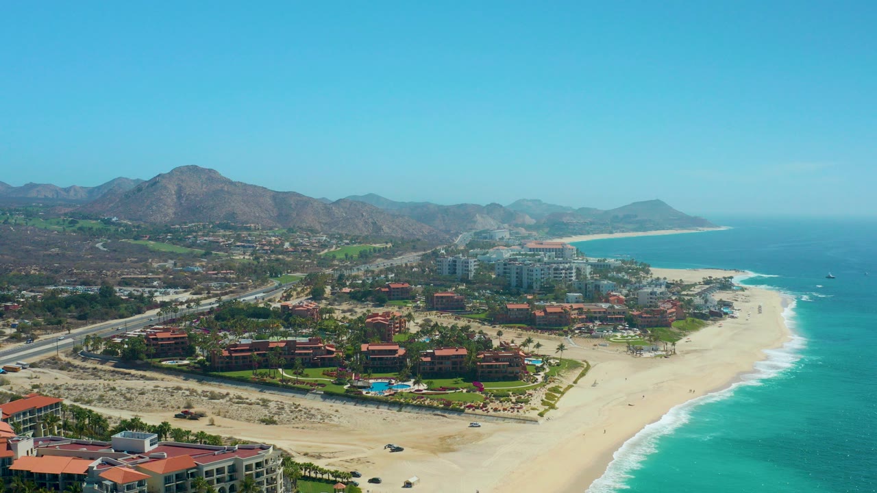 Aerial frame over Dreams Los Cabos showing red-roofed resort buildings beside a sandy beach, turquoise ocean, coastal highway, mountains, clear sky, and no visible people.