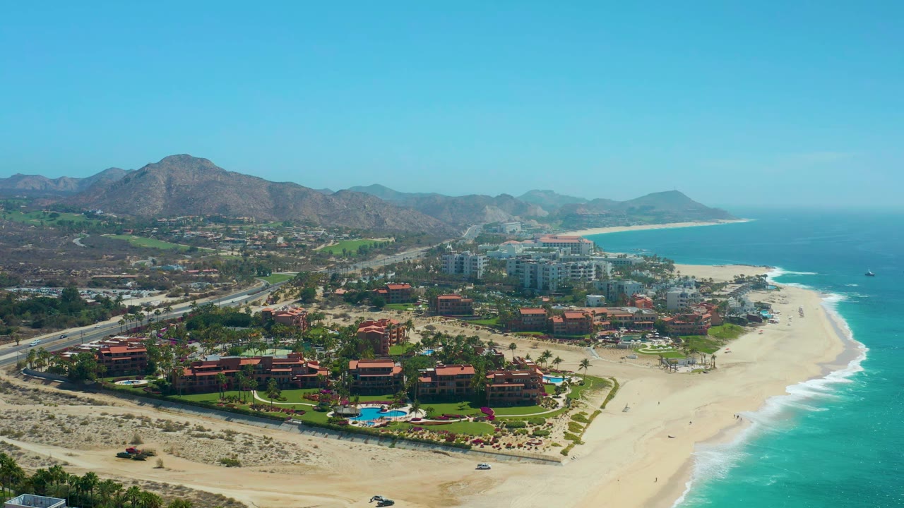 Drone view of resort buildings, beach, ocean waves, and a coastal highway.