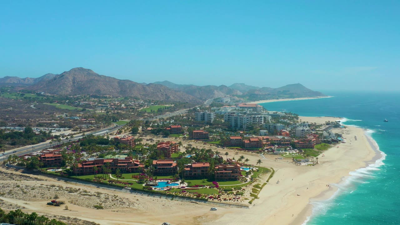 High drone frame over the Dreams Los Cabos beachfront, with resort towers near center, red-roof lodging in the lower frame, white surf tracing the sand, distant mountains, and tiny people on the beach.