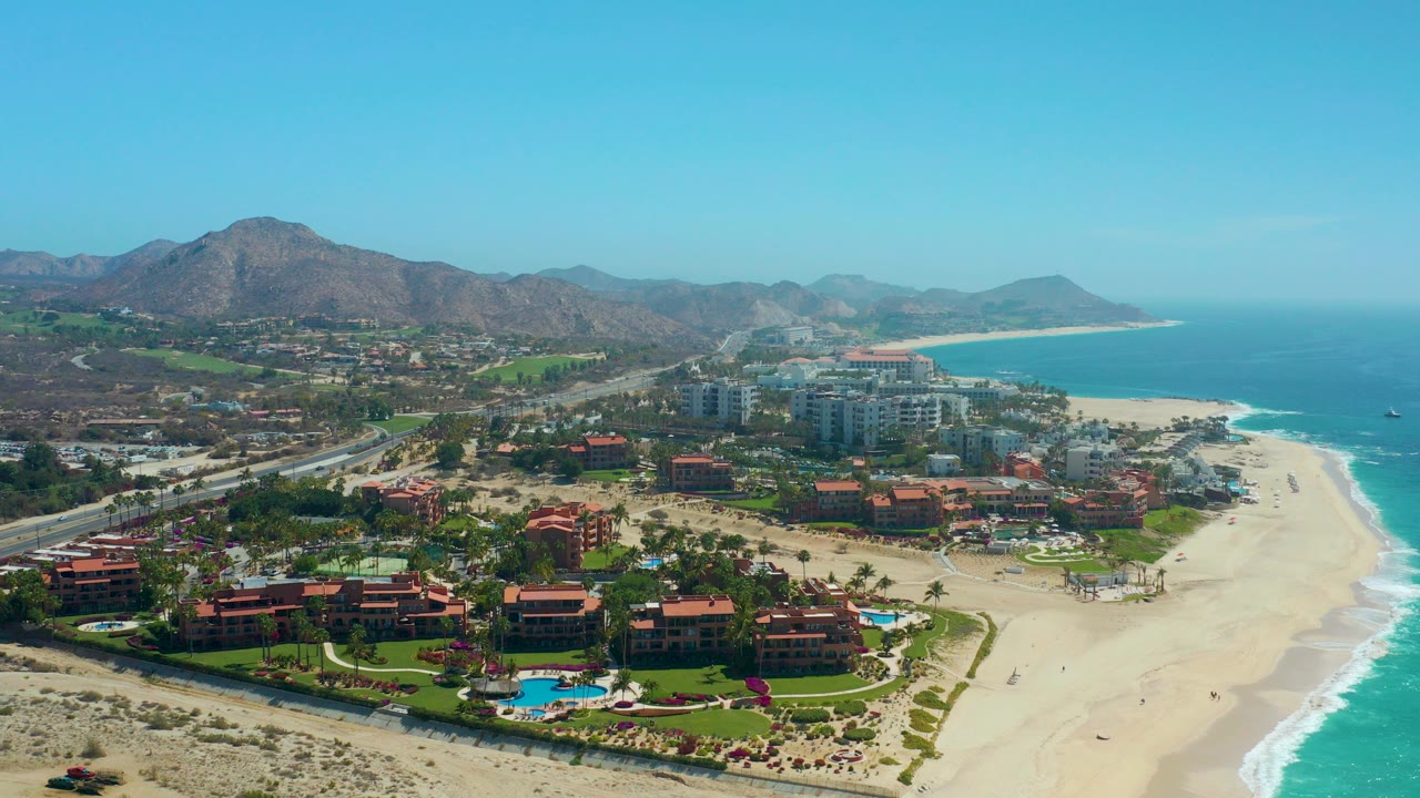 Opening aerial frame over Dreams Los Cabos in Baja California Sur, with red-roof resort buildings and pools below, a road to the left, pale beach and blue ocean to the right, and mountains on the horizon.