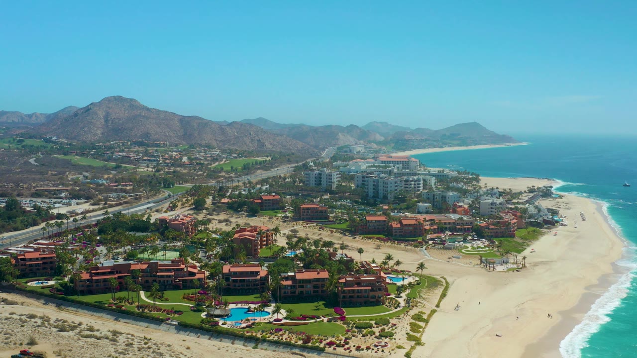 Aerial frame of Dreams Los Cabos with red-roof buildings and blue pools in the foreground, resort towers near the beach, a bright surf line on the right, and brown mountains beyond the developed coast.