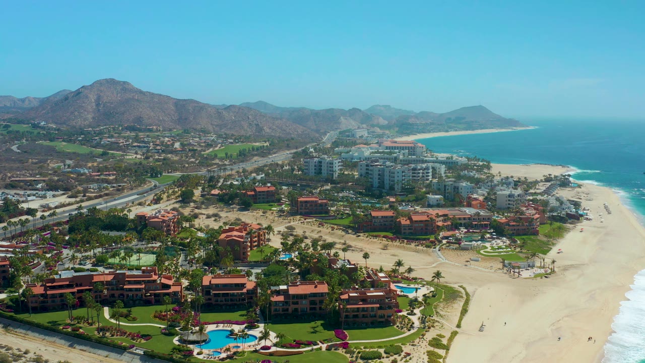 Aerial view over Dreams Los Cabos with resort lawns and pool in the foreground, hotel buildings and road beyond, open sand and ocean to the right, mountains in back, and no visible people.