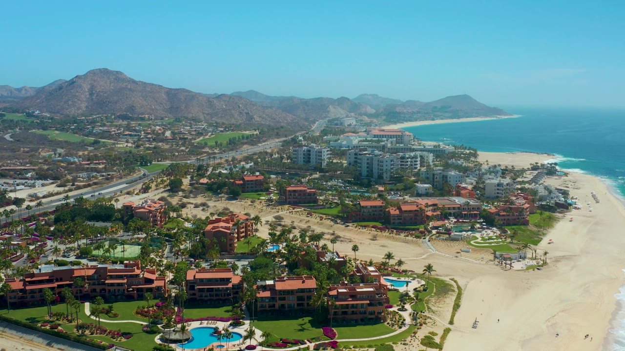 Wide aerial of resort pool, red roofs, sandy beach, ocean, and road