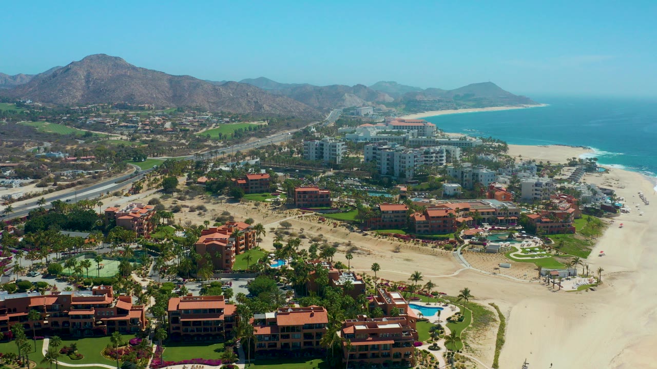 Wide aerial frame over Dreams Los Cabos showing red-roof resort buildings, pools, pale sand, blue ocean, and mountains at the start of the clip.