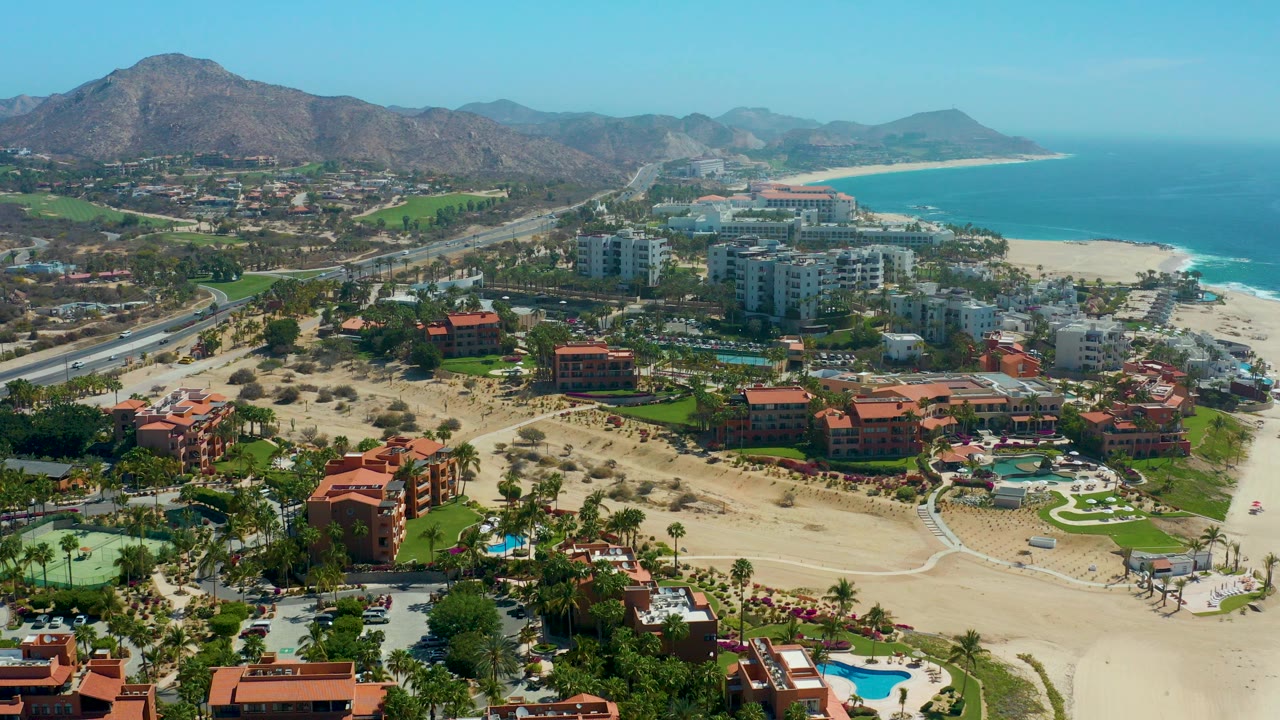 Wide aerial view of Dreams Los Cabos with red-roof buildings in the foreground, white high-rises near the beach, ocean to the right, road to the left, and mountains under a clear sky.