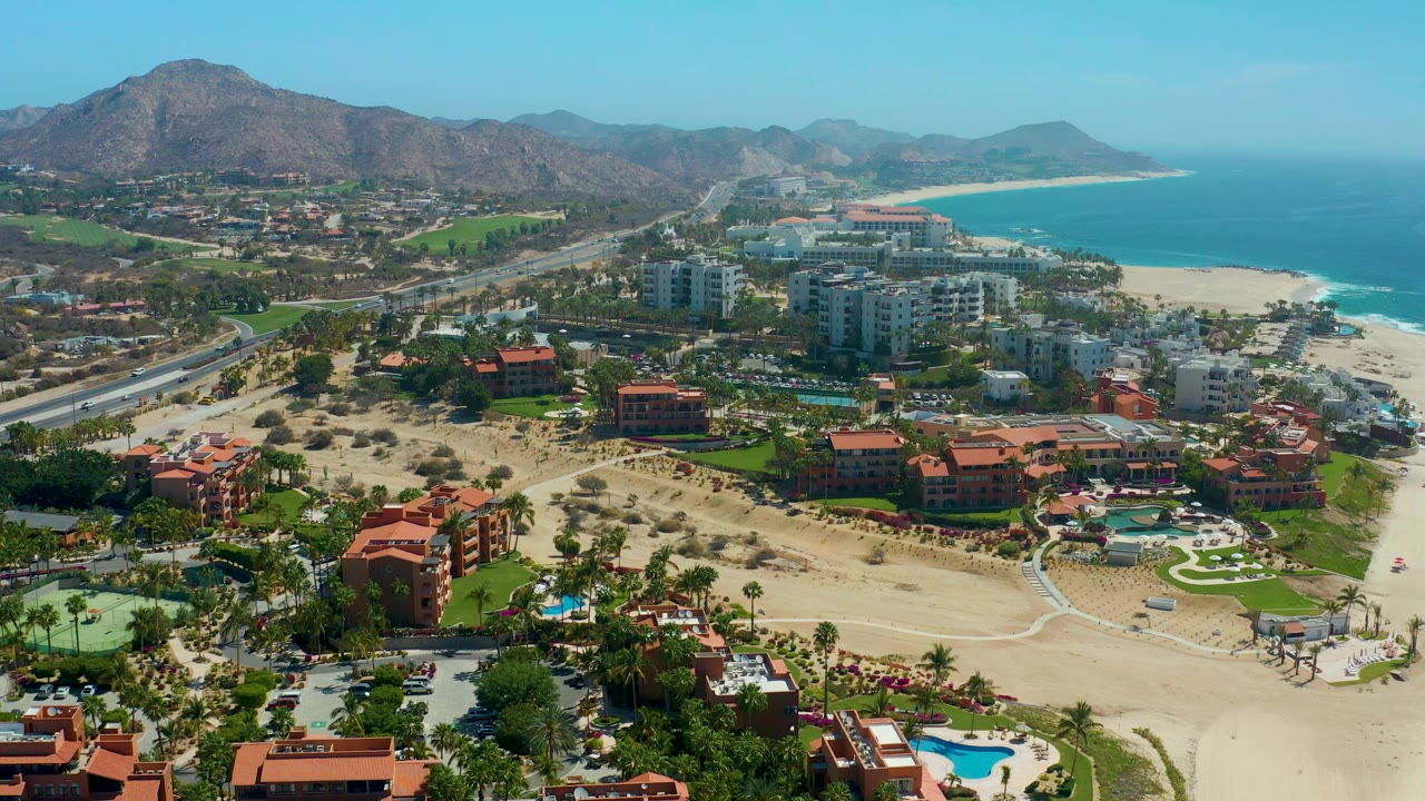 Aerial resort coastline with beach, road, high-rises, red roofs, ocean, and mountains.