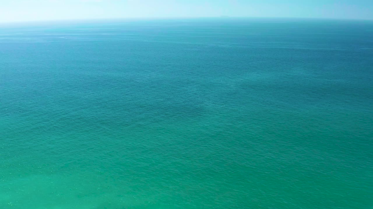 Aerial frame over calm turquoise water at Dreams Los Cabos, Mexico, showing a slightly deeper blue band near the horizon, faint seabed texture below, and no visible people.