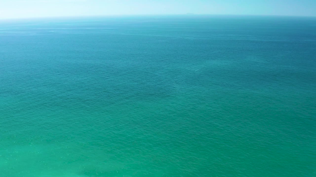 High aerial view of open water near Dreams Los Cabos, Mexico, with green shallows in the lower frame, deeper blue sea above, a hazy horizon, and no visible people.