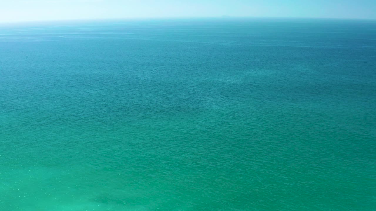 Mid-clip aerial view over clear blue-green ocean at Dreams Los Cabos, Mexico, with faint seabed shapes visible through the lower water and no visible people.