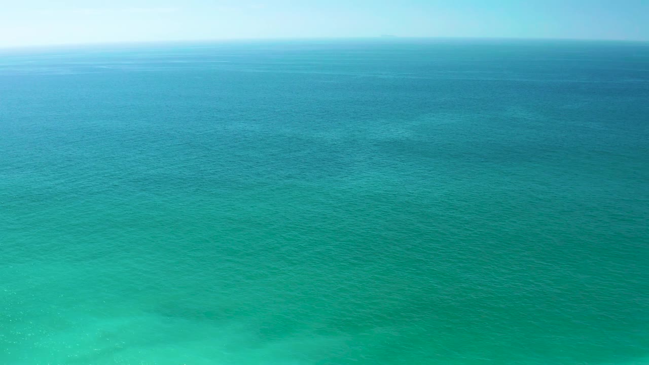 Aerial-looking frame over Dreams Los Cabos water, showing a broad turquoise surface, faint lighter patches near the lower frame, a level horizon, and no visible people.