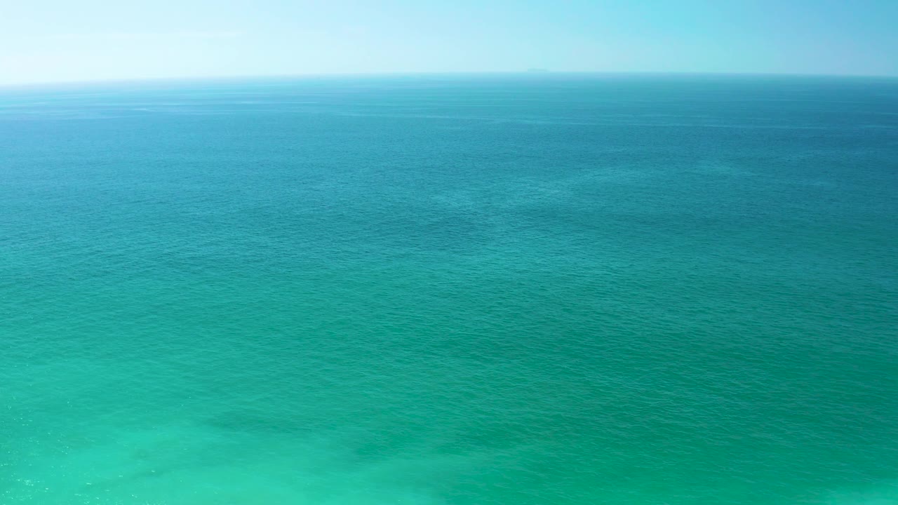 Mid-clip view of Dreams Los Cabos water, with bright turquoise shallows at the lower edge, darker blue ocean toward the horizon, pale sky, and no visible people.