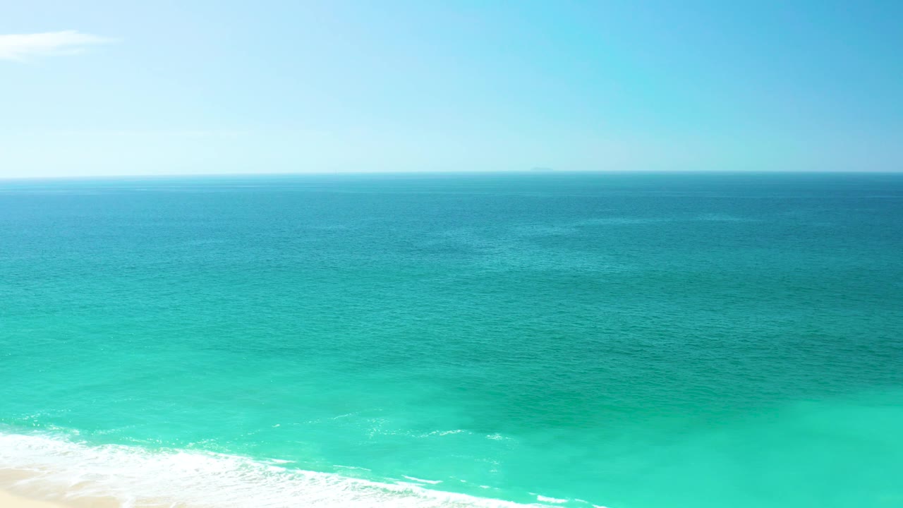 Midway through the clip, pale sand is more visible along the lower edge beneath turquoise water, while the blue horizon remains clear and no people are visible.