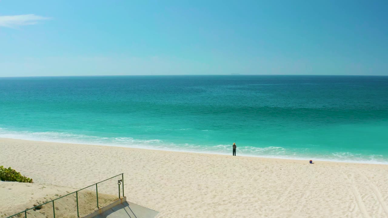 At Dreams Los Cabos in Baja California Sur, a wide elevated view shows turquoise ocean beyond pale sand, a foreground railing and stairs, and one distant person near the shore.