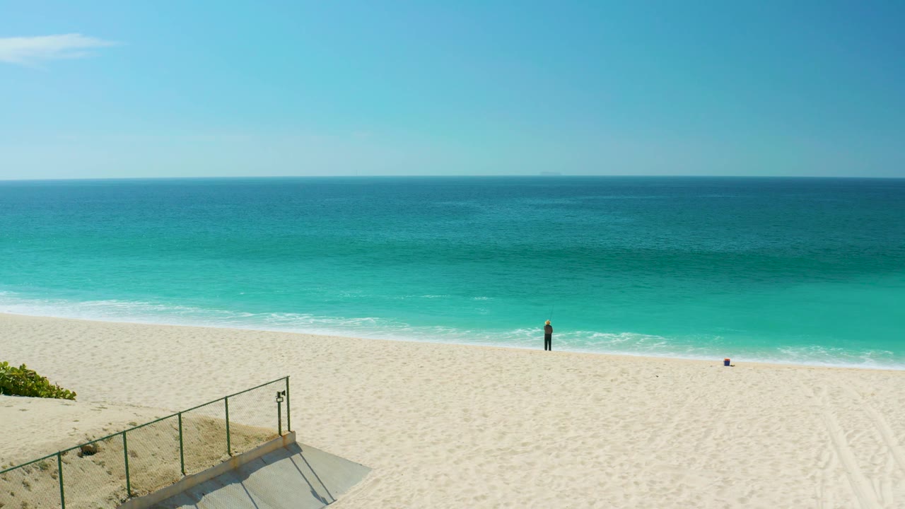 A sunlit Dreams Los Cabos beach frame shows the foreground railing angled across the lower edge, pale sand through the center, turquoise surf beyond, and one small person near the waterline.
