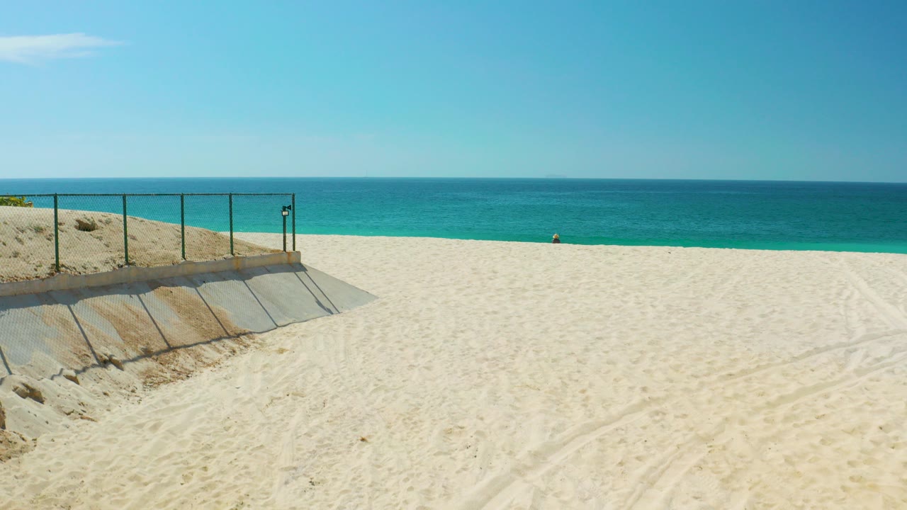 At Dreams Los Cabos in Baja California Sur, a wide beach frame shows pale sand, a concrete ramp with metal railing on the left, blue ocean on the horizon, clear sky, and no visible people.