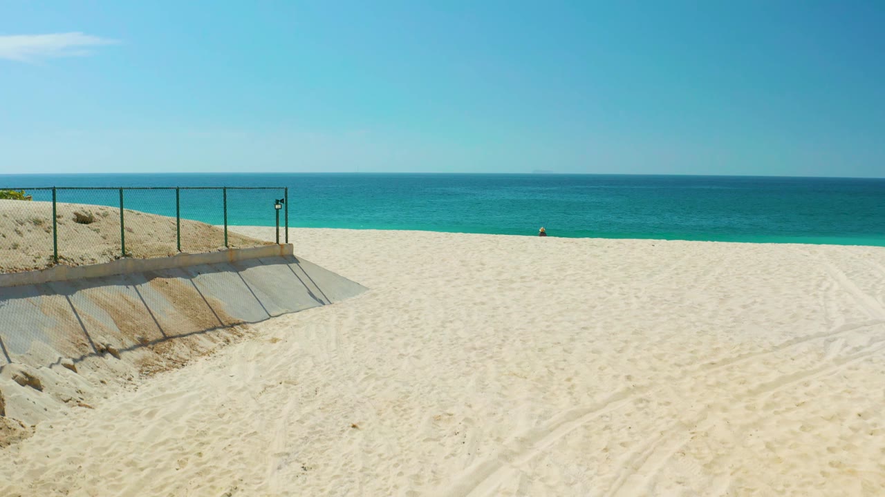 At Dreams Los Cabos in Baja California Sur, the beach view holds on the left-side concrete ramp and railing, with pale sand across the foreground, turquoise water beyond, and no visible people.
