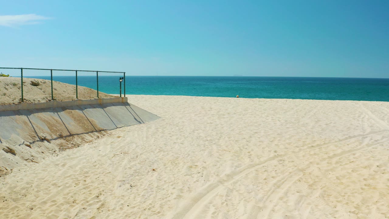 At Dreams Los Cabos in Baja California Sur, the frame shows the concrete ramp and fence line higher at left, broad empty sand in front, a blue ocean band in back, and no visible people.