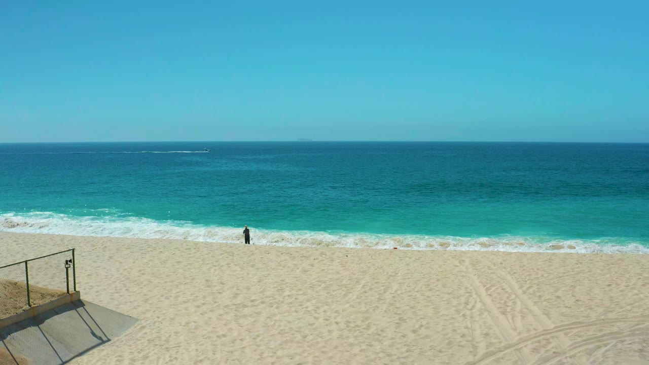 Opening frame at Dreams Los Cabos shows pale sand, turquoise waves, a clear blue sky, a small person by the surf, and a partial beach access railing at lower left.