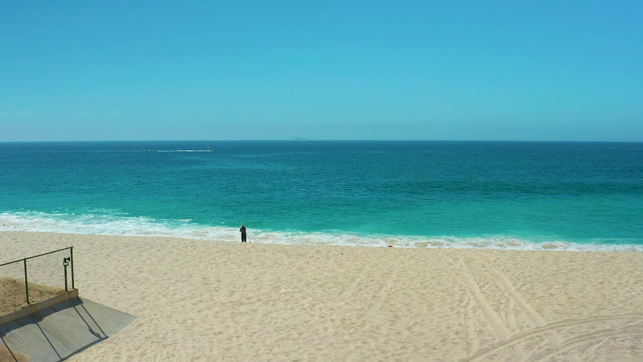 A wide Dreams Los Cabos beach frame shows the person still near the breaking waves, with tire-like sand tracks, open turquoise water, and a level blue horizon.