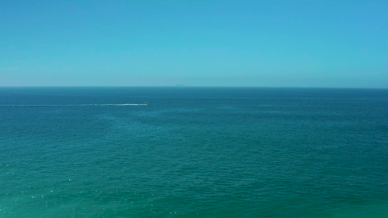 Wide aerial frame at Dreams Los Cabos with a small boat farther across the blue-green ocean, its white wake stretching behind it below a cloudless sky and no visible people.