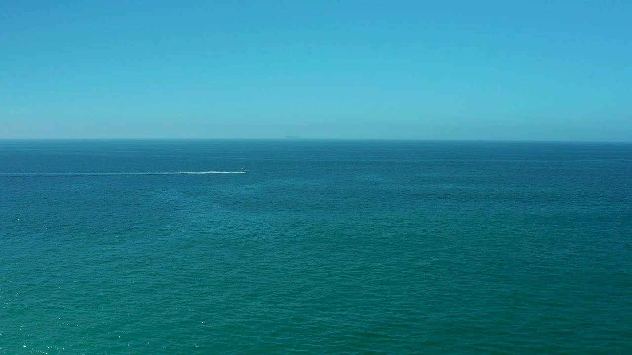 Aerial Los Cabos ocean view with the boat and bright wake positioned toward the right side of the horizon band, over calm turquoise and deeper blue water with no visible people.