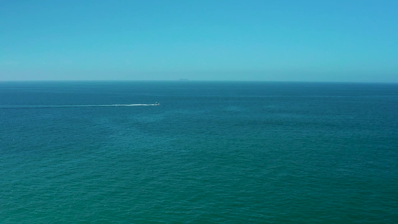Wide elevated view from Dreams Los Cabos showing open ocean, a faint distant shoreline or haze on the horizon, and a small boat wake crossing the upper frame with no visible people.