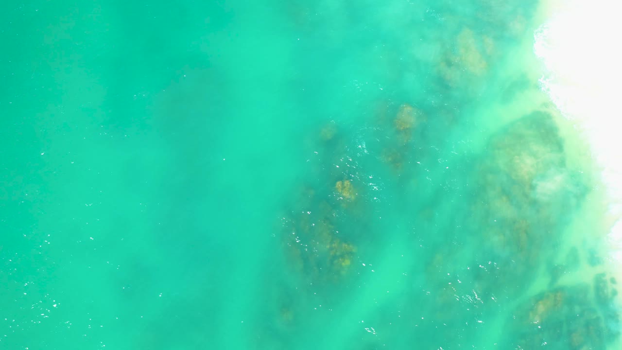Aerial view at Dreams Los Cabos shows translucent turquoise water, a darker rocky patch near center-right, pale wash at the upper-right edge, and no visible people.