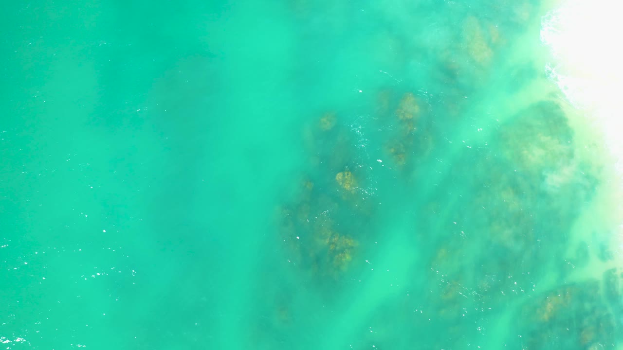 At Dreams Los Cabos, blue-green shallow water covers scattered brown rock clusters, with a brighter frothy area along the upper-right corner and no visible people.
