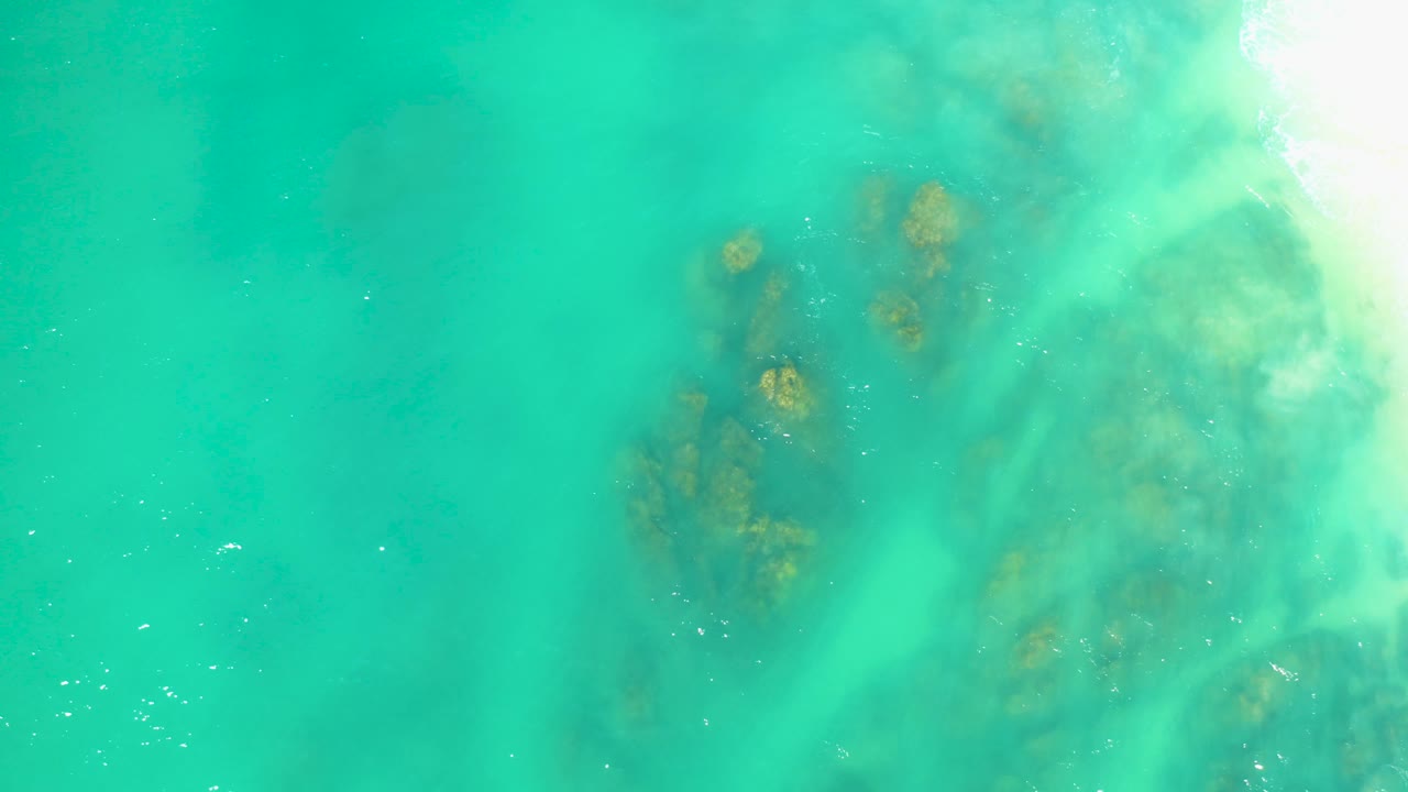 A top-down frame from Dreams Los Cabos shows a larger rocky area under turquoise water, with pale streaks of moving wash passing over it and no visible people.