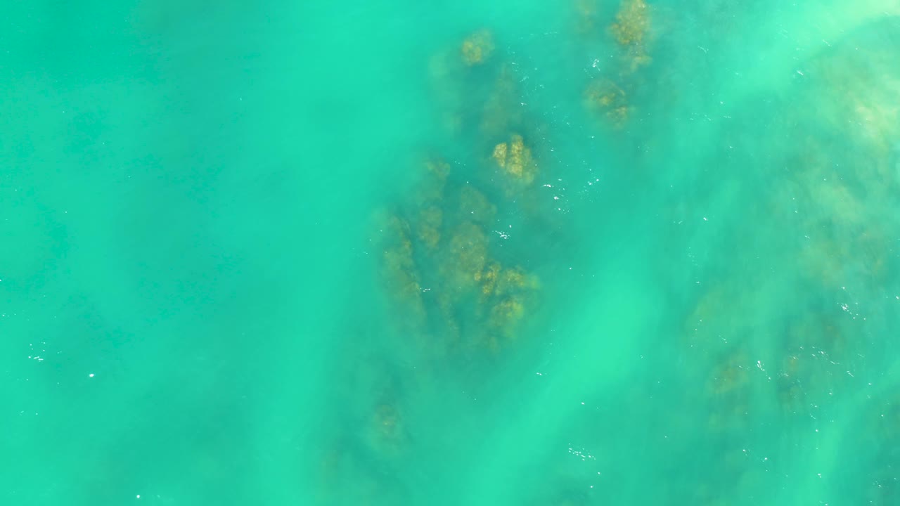 Turquoise ocean water at Dreams Los Cabos covers scattered dark rock or coral forms, with softer green seabed tones to the right and no visible people.