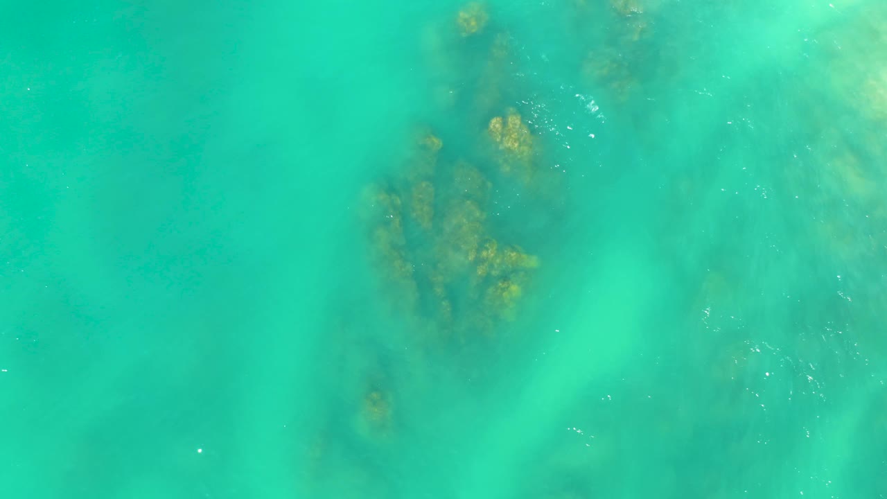 Shallow green-blue water at Dreams Los Cabos reveals a darker underwater formation near the center, pale seabed haze, and no visible people.